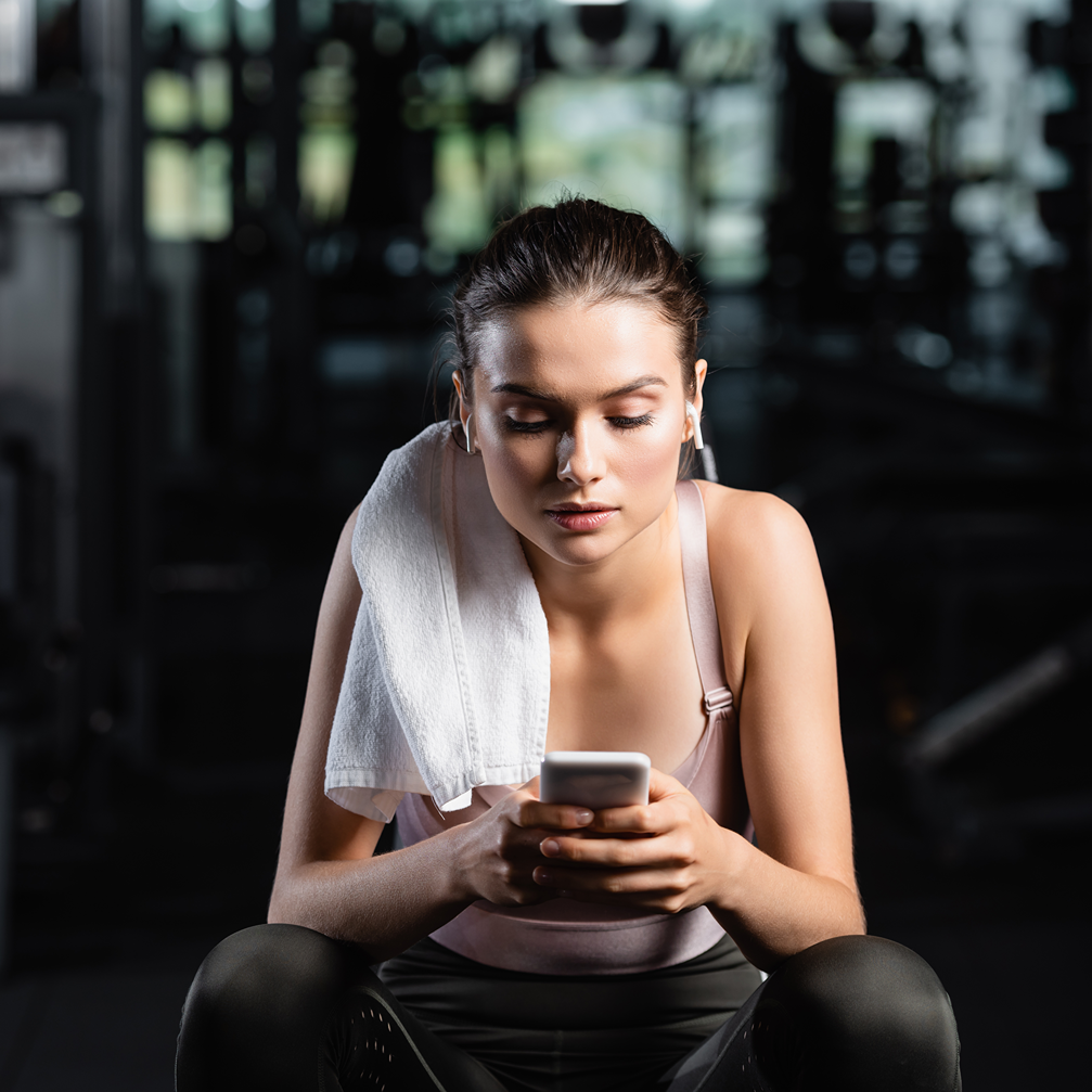 Eine Frau in Sportkleidung sitzt in einem Fitnessstudio, trägt ein Handtuch über der Schulter, nutzt ein Smartphone und hat kabellose Ohrhörer im Ohr.