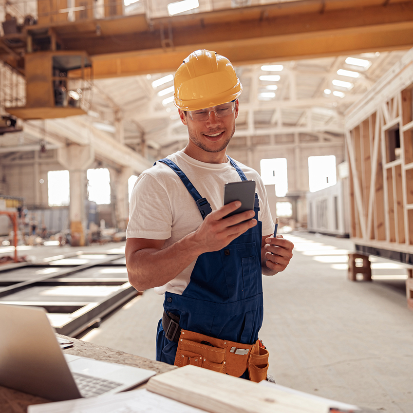 Ein lächelnder Arbeiter in blauer Arbeitskleidung und gelbem Schutzhelm steht in einer großen Werkhalle, hält ein Smartphone in der Hand und hat Werkzeug an seinem Gürtel.